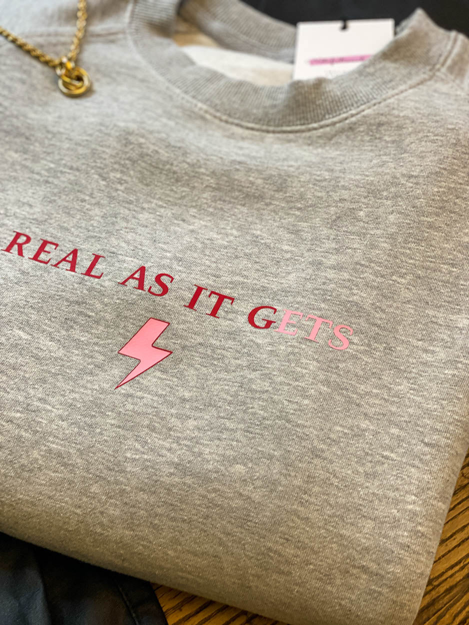 Grey-sweatshirt-close-up-slogan-Real-as-it-gets-with-pink-thunderbolt-detail